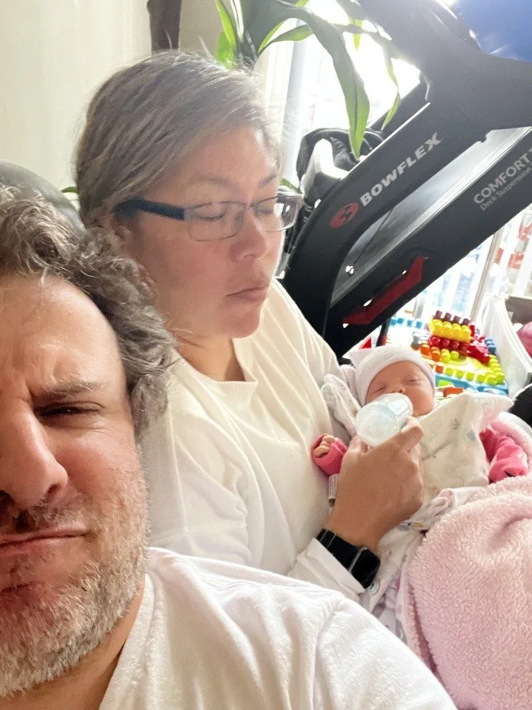 Paul in the foreground with Lina behind him feeding their newborn a bottle, in the early days before Suddenly Clean