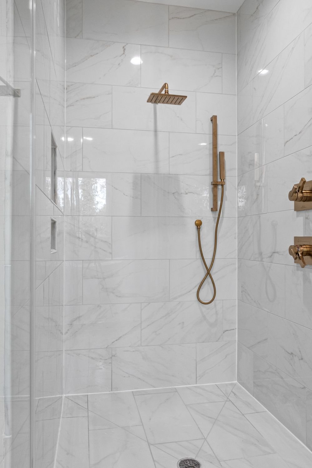 Walk-in shower with white marble-look tile and a brushed bronze rain shower head and hand shower
